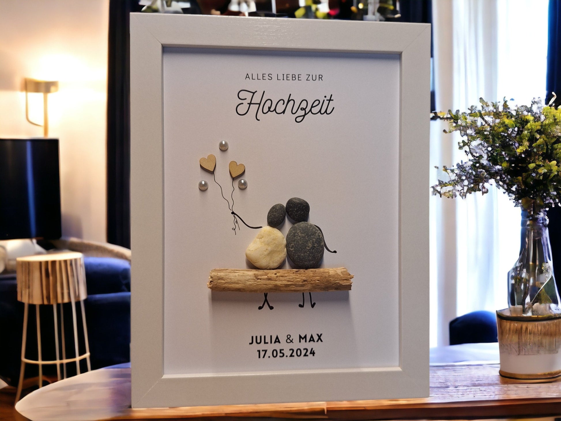 Steinbild bestehend aus dem Text Alles Liebe zur Hochzeit oberhalb der Steinfiguren, mittig im Bild sind 2 Steinfiguren (eine in weiß die andere in dunkel) auf einer Bank aus einem Treibholzbank platziert. Die linke Figur hat 2 Holzherzchenluftballons in der Hand, welche mit Perlen dekorier sind. Unterhalb der Steinfiguren wird der Name der Eheleute sowie das Hochzeitsdatum mittig gedruckt.