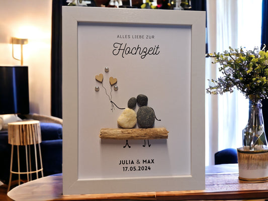Steinbild bestehend aus dem Text Alles Liebe zur Hochzeit oberhalb der Steinfiguren, mittig im Bild sind 2 Steinfiguren (eine in weiß die andere in dunkel) auf einer Bank aus einem Treibholzbank platziert. Die linke Figur hat 2 Holzherzchenluftballons in der Hand, welche mit Perlen dekorier sind. Unterhalb der Steinfiguren wird der Name der Eheleute sowie das Hochzeitsdatum mittig gedruckt.