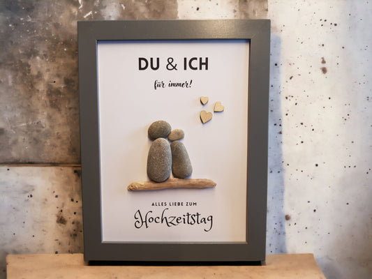 Steinbild mit dem Text du & ich für immer. Alles liebe zum Hochzeitstag. Abgebildet sind 2 Steinfiguren auf einem Treibholz, aneinandergeschmiegt und rechts oben 3 Holzherzen. Im Bilderrahmenfarbe nach Wahl. Hintergrund weißes Papier.